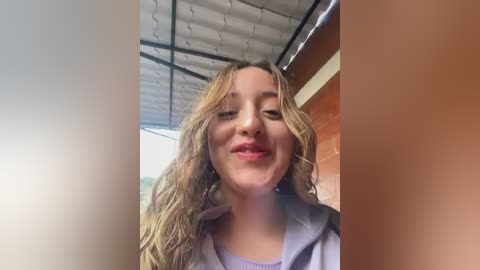 Video of a smiling, fair-skinned young woman with wavy, shoulder-length blonde hair, wearing a light purple hoodie, standing under a corrugated metal roof with an orange wall in the background.
