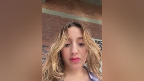Video of a young woman with wavy, shoulder-length blonde hair, fair skin, and light makeup, standing in front of a brick wall with graffiti, wearing a purple top.