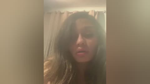 A blurry video of a woman with long, wavy hair, slightly tousled, wearing a light-colored top. She has a neutral facial expression, possibly in a dimly lit room with beige curtains in the background.