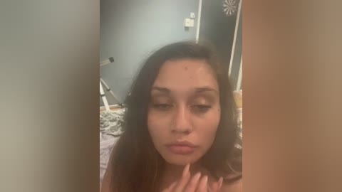 Video of a young Asian woman with long, straight brown hair, light skin, and closed eyes, standing in a dimly lit bedroom with a gray wall and a bed in the background.