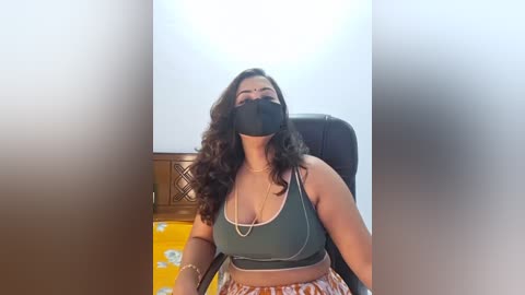 Video of a South Asian woman with long, wavy hair, wearing a green sports bra, orange and white shorts, and a black face mask. She sits in a chair with a wooden headrest, in a brightly lit room.