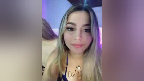 Video of a young woman with long, straight blonde hair, fair skin, and brown eyes, wearing a blue bra, taken indoors with a purple light background.