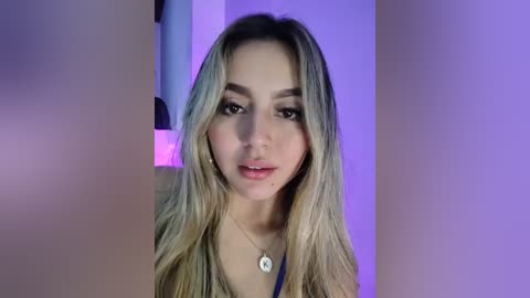 Video of a young, fair-skinned woman with long, wavy blonde hair and light makeup, wearing a blue top and a necklace. Background features purple lighting and a modern, minimalist room.