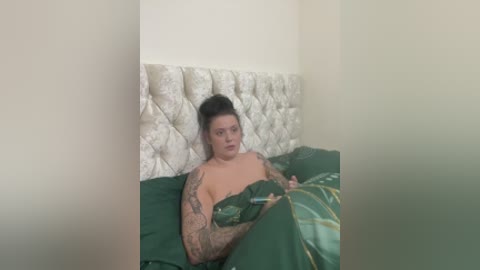 A video captures a tattooed woman with dark hair in a bun, lying on a green bed with a white, tufted headboard. She's partially covered by a green sheet, holding a glass, in a simple, intimate setting.