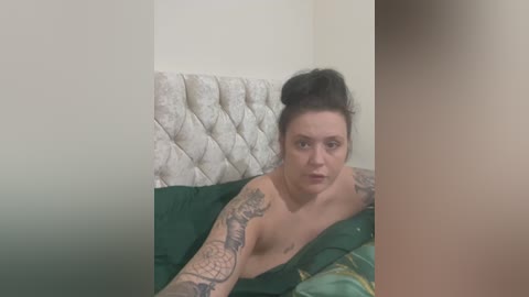 A video of a topless, light-skinned woman with tattoos and dark hair in a messy bun, lying on a bed with green sheets and a tufted white headboard.