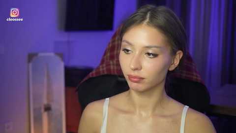 A video of a young Asian woman with light skin, brown hair, and delicate features, wearing a white tank top, sitting in a gaming chair with a red and black plaid pattern.