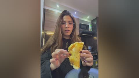 Video of a young woman with long brown hair, wearing a black sweater, holding a slice of pizza in a modern kitchen with wooden cabinets, under recessed lighting.