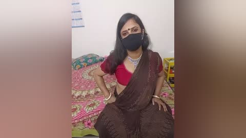Video of a South Asian woman with medium brown skin, wearing a maroon saree, red blouse, black face mask, and silver jewelry, seated on a bed with floral-patterned sheets.