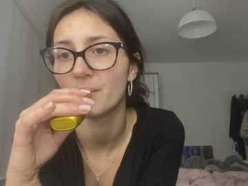 Video of a young woman with glasses, light skin, brown hair, and a black top, eating from a yellow container in a cluttered bedroom.