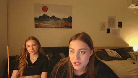Video of two young women, one with long brown hair, wearing a black top, in a cozy bedroom with a bed, framed art, and a warm light.