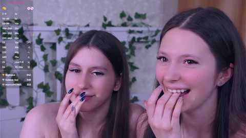 Video of two young women with long brown hair, one with blue nail polish, eating pizza, in a cozy indoor setting with hanging plants and a white background.