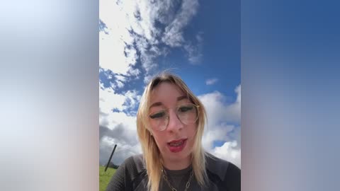 Video of a blonde woman with glasses, wearing a grey shirt, standing in a grassy field under a partly cloudy blue sky.