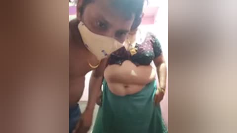 Video of a topless woman with medium skin tone, wearing a green skirt and a white face mask, partially covering her face. She has a belly and is in a doorway.
