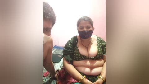 Video of a South Asian woman with dark skin, wearing a green sari with a pattern, black mask, and glasses. She has medium-sized breasts, sitting on a patterned bed in a pink-walled room.
