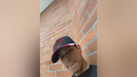 Video of a young Hispanic man with short hair, wearing a black cap with red logo and a black t-shirt, leaning against a red brick wall.
