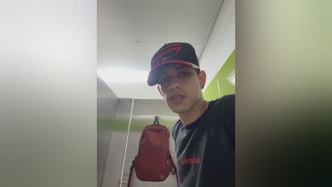 A video of a young man with light skin and short black hair, wearing a black cap and shirt, standing in a dimly lit bathroom with a red backpack, white walls, and a green door.