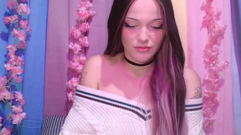 Video of a young woman with long, straight brown hair and a light complexion, wearing a white off-shoulder knit sweater, black choker, and pink floral backdrop.