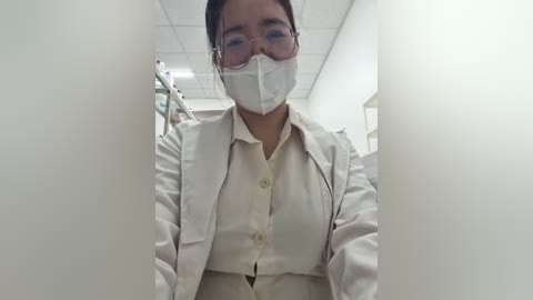 Video of an Asian woman with medium build and dark hair in a bun, wearing a white lab coat, mask, and glasses, standing in a sterile, white-walled laboratory.