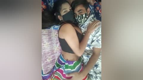 Video of a young woman with long dark hair, wearing a black bra and colorful patterned skirt, hugging a masked man in a white shirt, lying on a bed with colorful floral sheets.