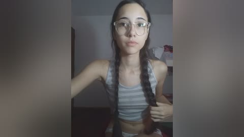 Video of a young woman with long dark hair in braids, wearing glasses and a striped crop top, standing indoors with blurred background.