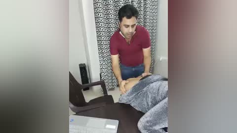 Video of a man with short dark hair in a maroon shirt, leaning over a man in a gray shirt, touching his stomach, in a room with a white curtain and black patterned drapes.