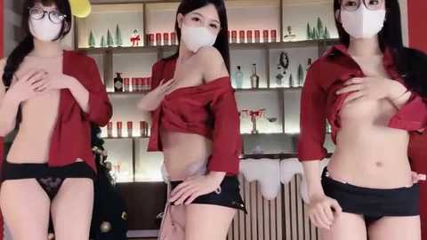 Video of three Asian women, all masked, topless, wearing red blouses, black shorts, and glasses, posing in a modern, well-lit room with shelves filled with bottles.