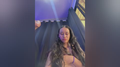 Video of a young woman with long, wavy black hair, partially nude, standing in a dimly lit room with black curtains, wearing a purple bra.