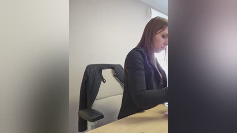 A video captures a woman with long brown hair, wearing a black jacket, seated at a wooden desk in a minimalist office with beige walls and a black chair.