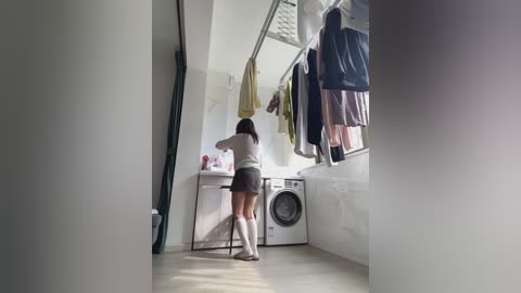 Video of a woman in a white top, gray shorts, and white socks doing laundry in a modern, brightly lit laundry room. Clothes hang on a drying rack, and a washing machine is visible in the background.