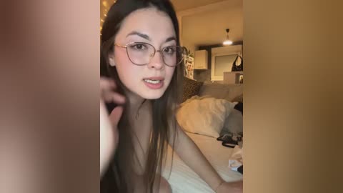 A video of an Asian woman with long black hair, wearing glasses, and light makeup, in a dimly lit bedroom, with a bed in the background.