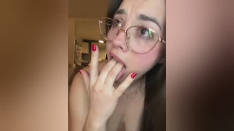 A close-up video of a young woman with fair skin, wearing large glasses and red nail polish, licking her finger suggestively, with a blurred, dimly lit background.