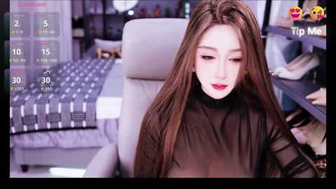 Video of an Asian woman with long brown hair, wearing a sheer black top, sitting on a white chair in a modern bedroom with a bed and shelves in the background.