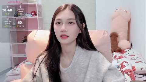A video of an East Asian woman with long black hair, wearing a gray sweater, sitting on a bed with a teddy bear. Background includes pink shelves and a soft pink wall.