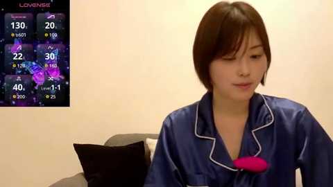 Video of a young woman with short brown hair wearing a blue satin pajama set with a pink heart-shaped pillow. Background includes a TV screen displaying a weather forecast.