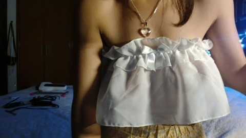 Video of a young woman with light skin, wearing a white, strapless top with ruffled edges and a gold necklace with a heart pendant. Background shows a dimly lit room with a bed and wooden wardrobe.