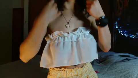 Video of a young woman in a dimly lit room, wearing a white, ruffled crop top and yellow shorts, with a black watch on her left wrist.