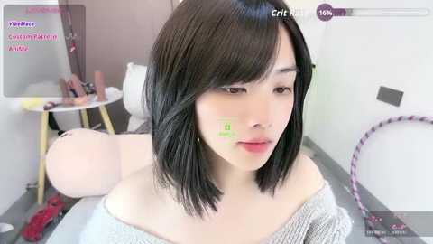 Video of a young East Asian woman with straight black hair, wearing a gray off-shoulder sweater, sitting on a bed in a bedroom.