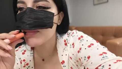 Video of a light-skinned woman with long black hair, wearing a floral blouse, black face mask, and orange lipstick, smiling, in a room with a brown couch and white walls.