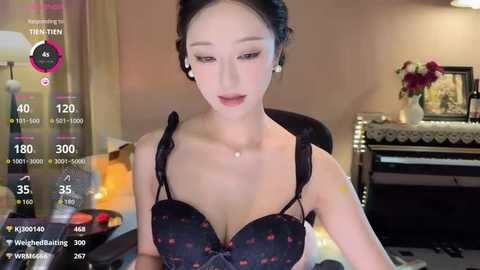 Media: A video of an East Asian woman with fair skin, wearing a black bra with pink floral designs, sitting in a dimly lit room.