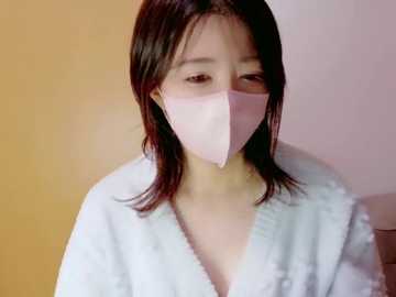 Video of a young Asian woman with shoulder-length dark hair, wearing a light pink surgical mask and a white hospital gown. The background is a gradient of light orange and beige.