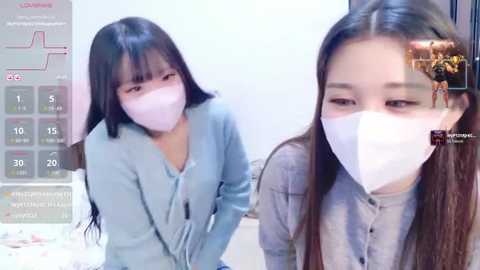 Video of two East Asian women with long black hair, wearing white masks and light blue sweaters, taken indoors with a video game interface overlay.