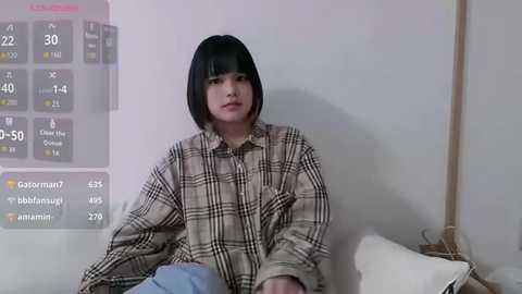 A young East Asian woman with a short black bob, wearing a beige plaid shirt, sits on a white couch, with a digital weather forecast overlay on the wall behind her.