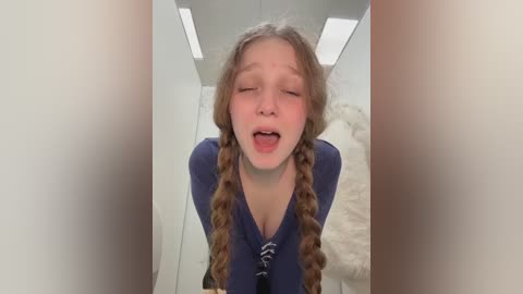 Video of a young girl with long, braided brown hair, wearing a blue dress, in a modern, minimalist room with white walls and a fluffy white rug.