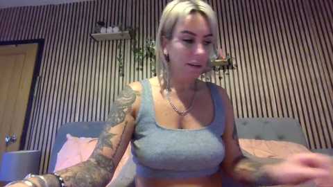 A video of a blonde woman with tattoos, wearing a grey crop top, seated on a bed with striped wallpaper. She has a confident expression and is interacting with something off-camera.