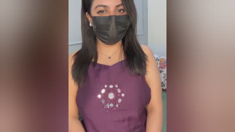 Video of a young South Asian woman with medium skin tone, wearing a black face mask, black hair, and a purple sleeveless shirt with a silver flower design, standing indoors.