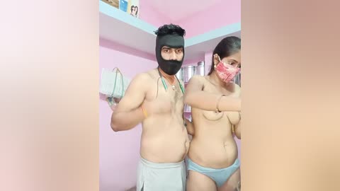 Video of two shirtless men with masks and light blue shorts, standing in a pink room with shelves and a blue ceiling, one covering his crotch, the other covering his chest.