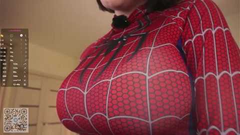 A close-up video of a woman in a red Spider-Man costume with large breasts, standing indoors next to a white door.