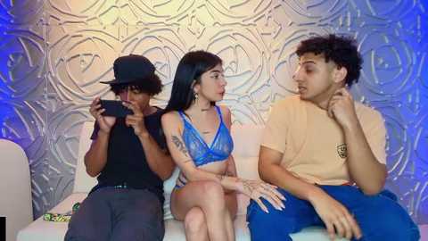 Video of three young people: a girl in a blue bra and panties, a boy in a beige shirt, and a person in a black hat taking a photo. Background: textured, ornate wall with blue lights.