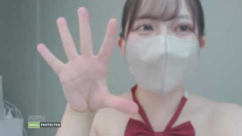 Video of an Asian woman with light skin and dark hair, wearing a red halter top and a white face mask, making a hand gesture against a blurred, light-colored background.