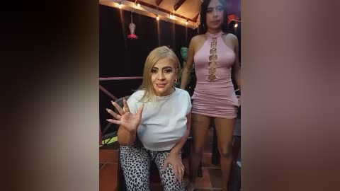 Video of a blonde woman in a white top and leopard print pants, gesturing, alongside a brunette in a pink dress, in a dimly lit room with string lights.